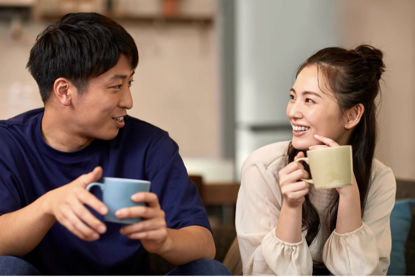 Two people sitting on a couch, with coffee mugs, talking and staring at each other.